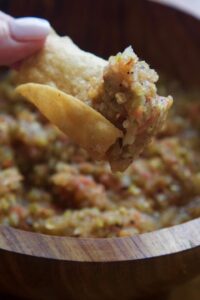Close-up of a hand holding a chip with salsa over a wooden bowl filled with chunky vegetarian green chile salsa.
