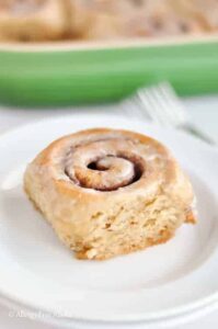 A cinnamon roll with icing on a white plate, accompanied by a green baking dish and a fork in the background, makes for the perfect addition to any tapioca flour recipes collection.