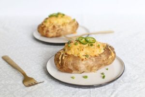 Two baked potatoes topped with cheese, jalapeño slices, and green onions on white plates with golden forks on a light patterned tablecloth—a perfect addition to your vegetarian green chile recipes.
