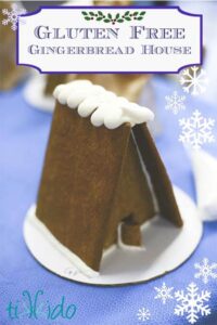 A small gluten-free gingerbread house, crafted with tapioca flour recipes and adorned with white icing on the roof, stands proudly against a blue background decorated with snowflakes.