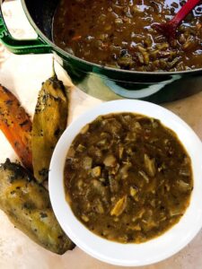 A bowl filled with green chili stew sits next to roasted green and red chili peppers, offering a delightful taste from popular vegetarian green chile recipes. A pot of additional stew and a ladle are in the background.