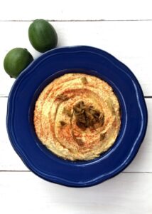 A blue plate filled with hummus, garnished with paprika and chopped olives, is accompanied by two limes on a white wooden surface, perfect for those seeking delicious vegetarian green chile recipes.
