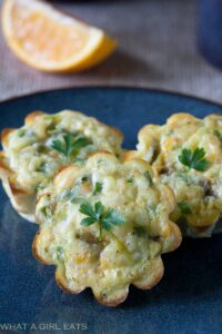 Three savory vegetarian muffins with herb garnishes are placed on a dark blue plate, accompanied by a slice of orange in the background, inspired by green chile recipes.