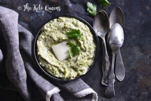 A bowl of mashed vegetables topped with a pat of butter, garnished with parsley, placed on a dark surface next to a folded gray napkin and three spoons is the perfect dish for those seeking delicious vegetarian keto recipes. "The Keto Queens" text appears in the top left.