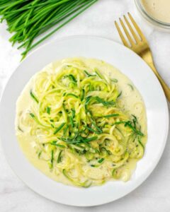 A plate of zucchini noodles topped with a creamy sauce and garnished with chopped chives, placed next to a gold fork on a white surface; an elegant dish perfect for vegetarian keto recipes enthusiasts.