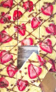 A tray of sliced dessert bars topped with strawberry slices, chocolate chips, and a chocolate drizzle. One bar is being lifted from the tray, perfect for those exploring vegetarian keto recipes.