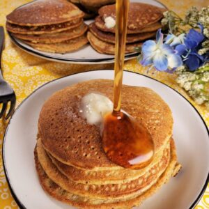 A stack of pancakes on a white plate with a dollop of butter on top and syrup being poured over them, reminiscent of indulgent vegetarian keto recipes. Another stack of pancakes and flowers adorn the background.