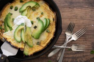 A skillet frittata, a star of vegetarian keto recipes, is topped with sliced avocado, a dollop of sour cream, and chopped herbs on a wooden table with a stack of four forks beside it.