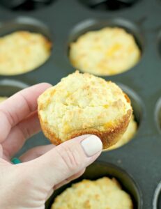 A hand holding a freshly baked muffin over a muffin tin containing more muffins, perfect for those exploring vegetarian keto recipes.