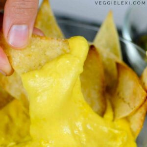 Close-up of a hand dipping a tortilla chip into a yellow cheese sauce. The cheese sauce is thick and creamy, coating the chip perfectly. Text in the top right corner reads "VEGGIELEXI.COM" featuring tapioca flour recipes.