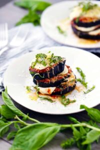 A plated dish of stacked eggplant slices with melted cheese, tomato sauce, and chopped basil, garnished with more basil leaves around the plate—a delightful addition to vegetarian keto recipes.