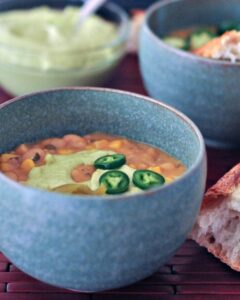 A blue bowl of soup topped with green sauce and sliced jalapenos, accompanied by a piece of bread on the side—an exquisite take on vegetarian green chile recipes. Another bowl of a similar dish graces the background, completing this culinary delight.
