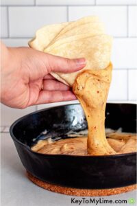 A hand is holding folded tortillas, dipping them into a skillet filled with melted, stringy cheese. The background features white tiled walls. For a twist, try using tapioca flour recipes to create gluten-free tortillas that are just as delicious and stretchy!