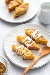 Two plates each hold three triangular banana rice cakes drizzled with coconut cream, accompanied by a wooden spoon. A small jug is placed nearby.