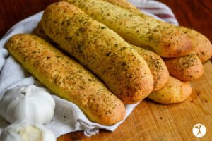 A bundle of seasoned breadsticks is stacked on a wooden surface next to two garlic bulbs and a white cloth, perfect for adding a delicious crunch to your vegetarian keto recipes.