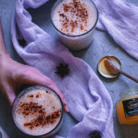 image of a hand holding a glass of eggnog next to a lavender colored gauzy cloth and a jar of cbd honey