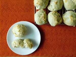 A white plate with two bread rolls sits on an orange surface next to a cooling rack holding several more bread rolls topped with sesame seeds, perfect for exploring your favorite tapioca flour recipes.