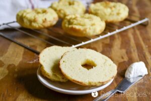 Two sliced bagels are placed on a small plate beside a knife with a dollop of cream cheese, perfect for those exploring vegetarian keto recipes. Four whole bagels are cooling on a wire rack in the background.