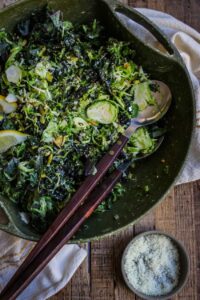A green ceramic bowl filled with a leafy green salad, garnished with lemon slices and seeds. Wooden-handled serving utensils rest in the bowl, with a small dish of grated cheese beside it—perfect for those exploring delicious vegetarian keto recipes.