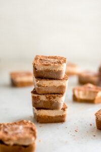 A stack of four cubed dessert slices with a brown topping, surrounded by scattered crumbs. Other similar dessert slices are in the background.