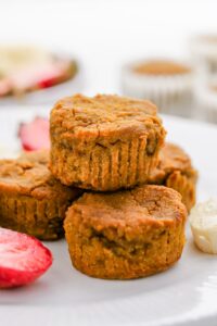 Four pumpkin muffins stacked on a white plate, with sliced strawberries and apple pieces nearby—perfect for those exploring tapioca flour recipes.