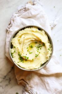 A bowl of creamy mashed potatoes garnished with chopped chives sits on a white cloth, perfect for those exploring vegetarian keto recipes.