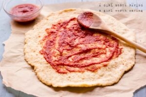 Pizza dough on parchment paper with tomato sauce spread on top; a wooden spoon with sauce and a small bowl of sauce are beside it, perfect for those exploring tapioca flour recipes.