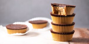 Stack of four chocolate peanut butter cups with a bite taken out of the top one, with two more cups on white paper liners in the background. Perfect for those seeking indulgent yet vegetarian keto recipes.