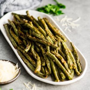 A rectangular white dish filled with roasted green beans, perfect for vegetarian keto recipes, is placed on a gray surface next to a small bowl of grated cheese and a sprig of parsley.