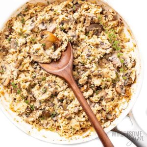 A skillet with creamy cauliflower rice and mushrooms, garnished with herbs, highlights the perfect blend of flavors in this vegetarian keto recipe, with a wooden spoon placed in the dish.