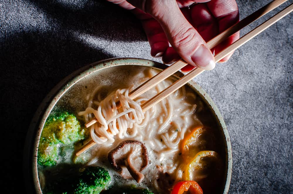 vegan ramen noodles in white bowl on gray background with a hand holding chopsticks