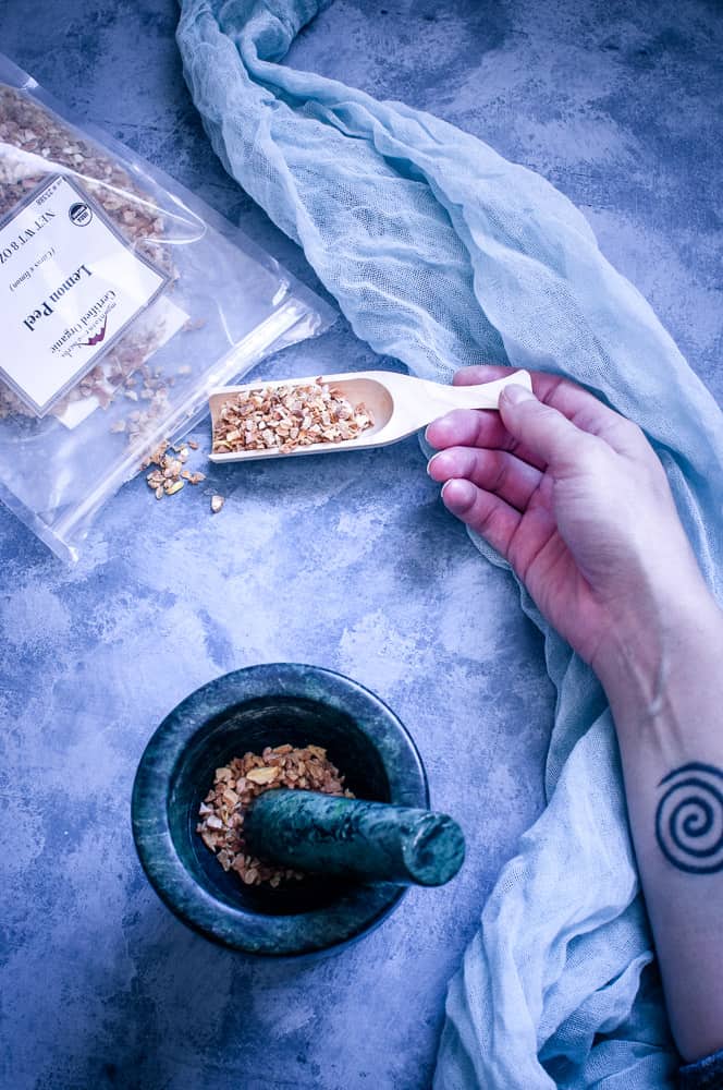 a top view flat lay of mountain rose herbs lemon peel spilled out next to mortar and pestle and arm with spiral tattoo holding wooden scoop