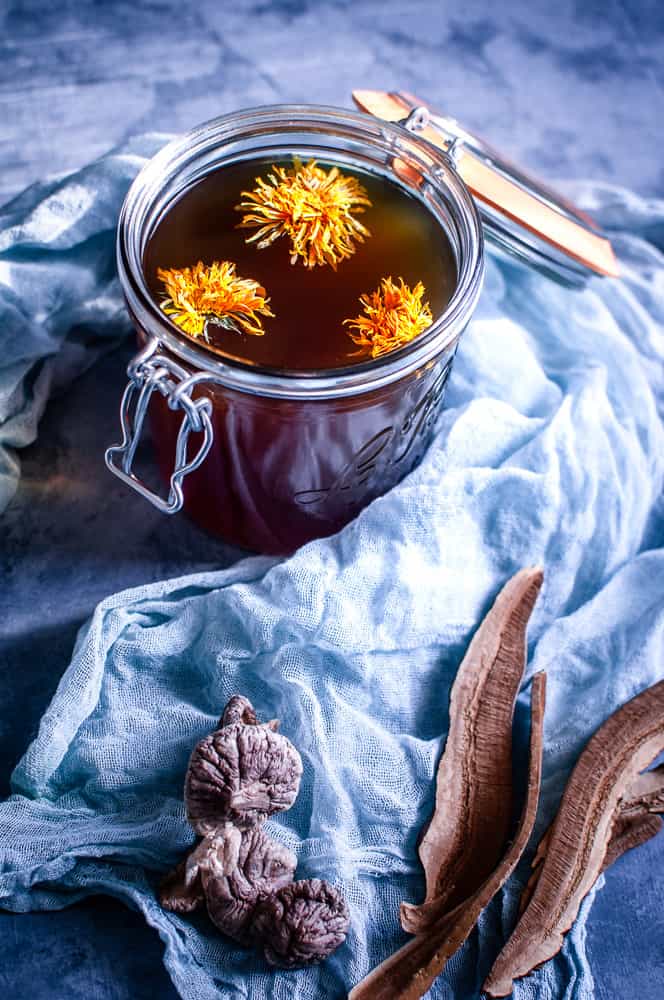 a clear jar filled with mushroom broth topped with edible flowers with mushrooms in the foreground on a sage gauze cheesecloth