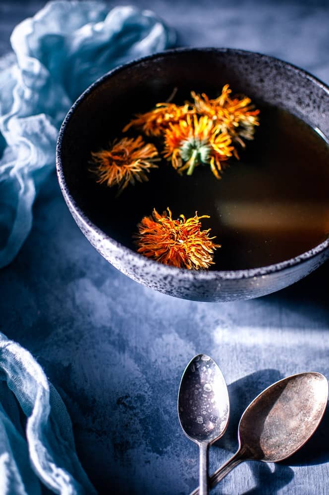 a gray backdrop with a black bowl filled with mushroom broth topped with calendula flowers alongside vintage spoons