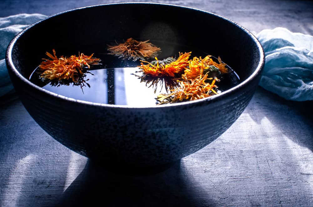 side shot view of black bowl filled with herbal broth topped with calendula flowers and the soft glow of light