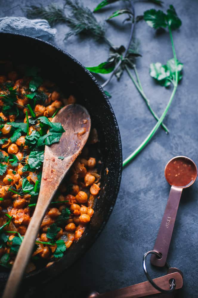 a cast iron skillet filled with spicy chickpeas topped with fresh herbs alongside a measuring spoon filled with harissa sauce