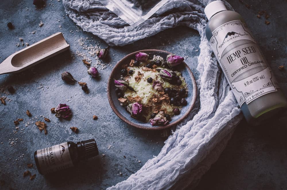 mountain rose herbs wooden scoop hemp oil and rosemary oil displayed next to a shallow bowl of DIY herbal bath salts