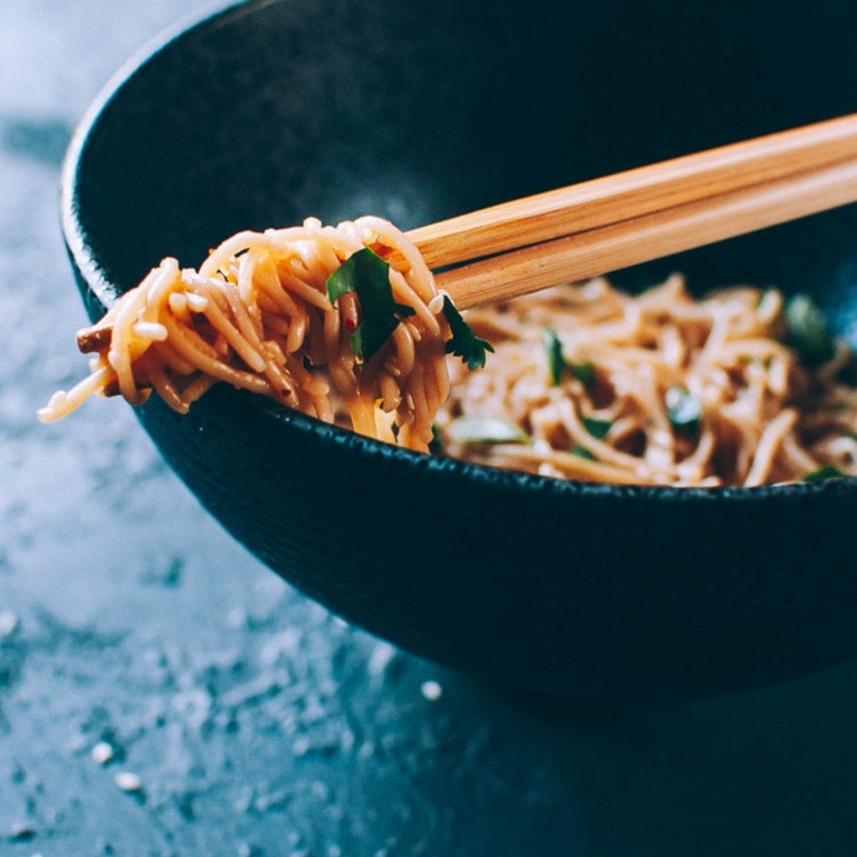Chopsticks lifting sesame noodles from a dark bowl filled with perfectly seasoned noodles, garnished with fresh herbs.