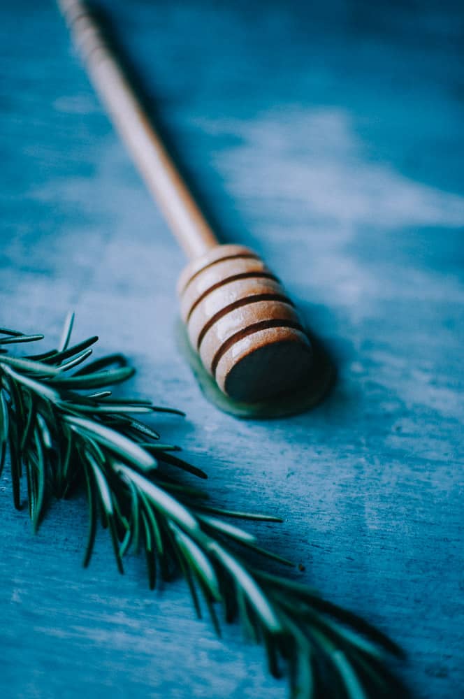 a wooden honey spoon dipped in honey laying next to a sprig of fresh rosemary