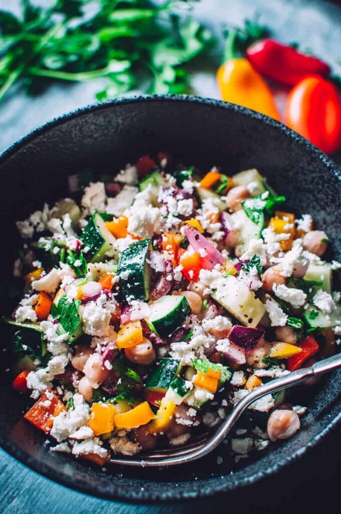 A silver spoon stuck in a black bowl of a salad with peppers, cucumber, feta cheese, red onion, chickpeas and more.