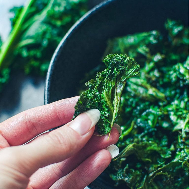 A hand holds a piece of crispy kale chip above a bowl brimming with more kale chips, freshly made in the air fryer.