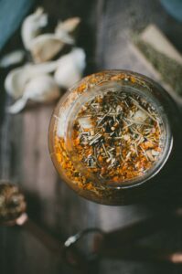 A glass jar filled with dried herbs and orange flower petals sits on a wooden surface, suggesting the beginnings of a fire cider recipe, with loose herbs and garlic cloves arranged in the background.
