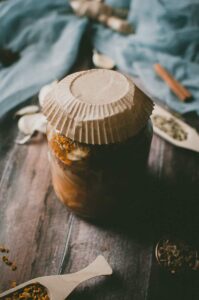 A glass jar covered with a paper lid contains preserved ingredients for a traditional fire cider recipe, sitting on a wooden surface with spices and a blue cloth in the background.