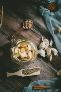 A jar with sliced ginger, surrounded by garlic cloves, dried herbs, a cinnamon stick, and measuring spoons on a wooden surface with a blue cloth—these ingredients are perfect for preparing a traditional fire cider recipe.