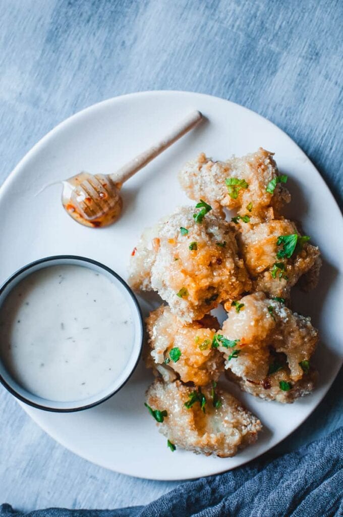 a white plate with a dish of ranch dressing and a honey spoon with habanero honey next to a pile of cauliflower wings