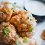 a close up shot of cauliflower wings sprinkled with parsely on a white plate next to a honey spoon