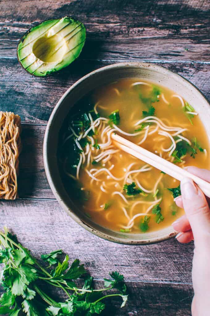 a hand holding chopsticks in a bowl of pho with ramen noodles