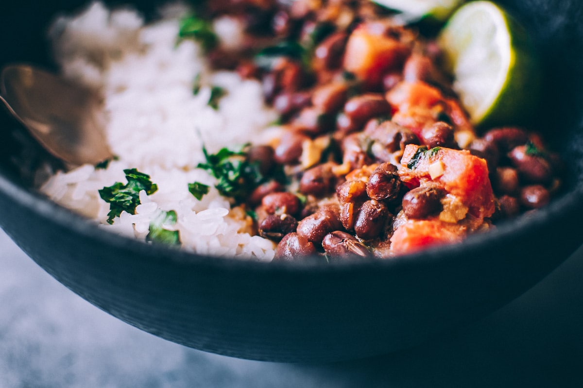 Close-up of a bowl with white rice, black beans, chopped herbs, and a lime wedge&mdash;perfectly complementing a side of Adzuki Bean Curry. A metal spoon is partially visible on the left side.