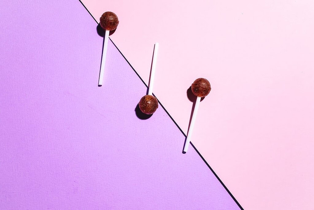 Three brown lollipops on white sticks are arranged diagonally on a background split into light pink and purple sections, offering a fun and colorful display of healthy candy for kids.