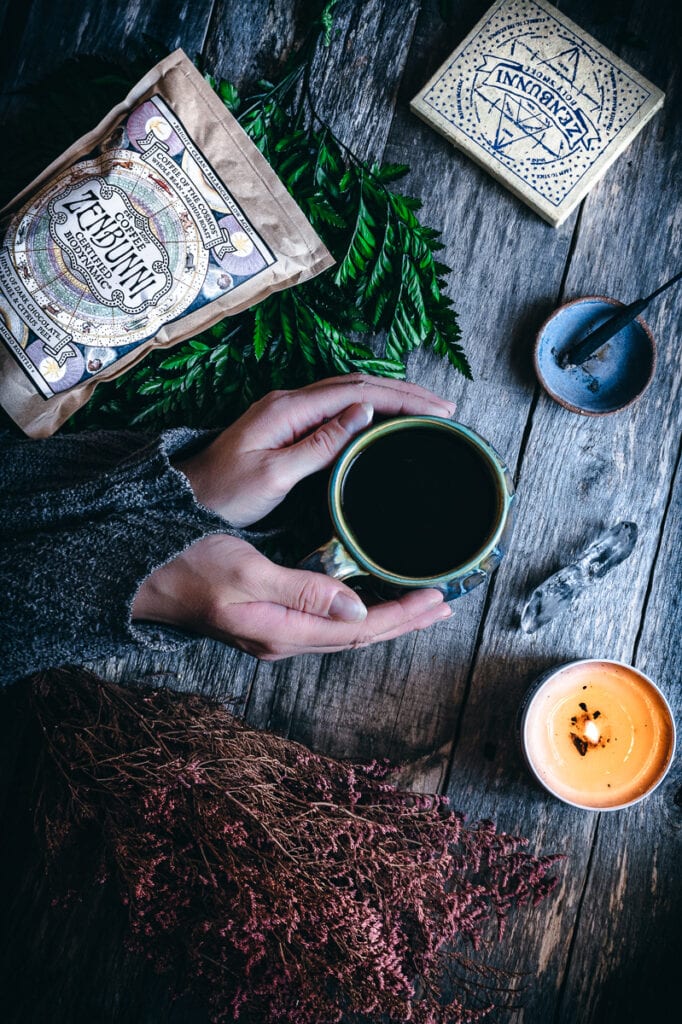 a flatlay of hands holding a coffee cup on a wooden table next to lit incense